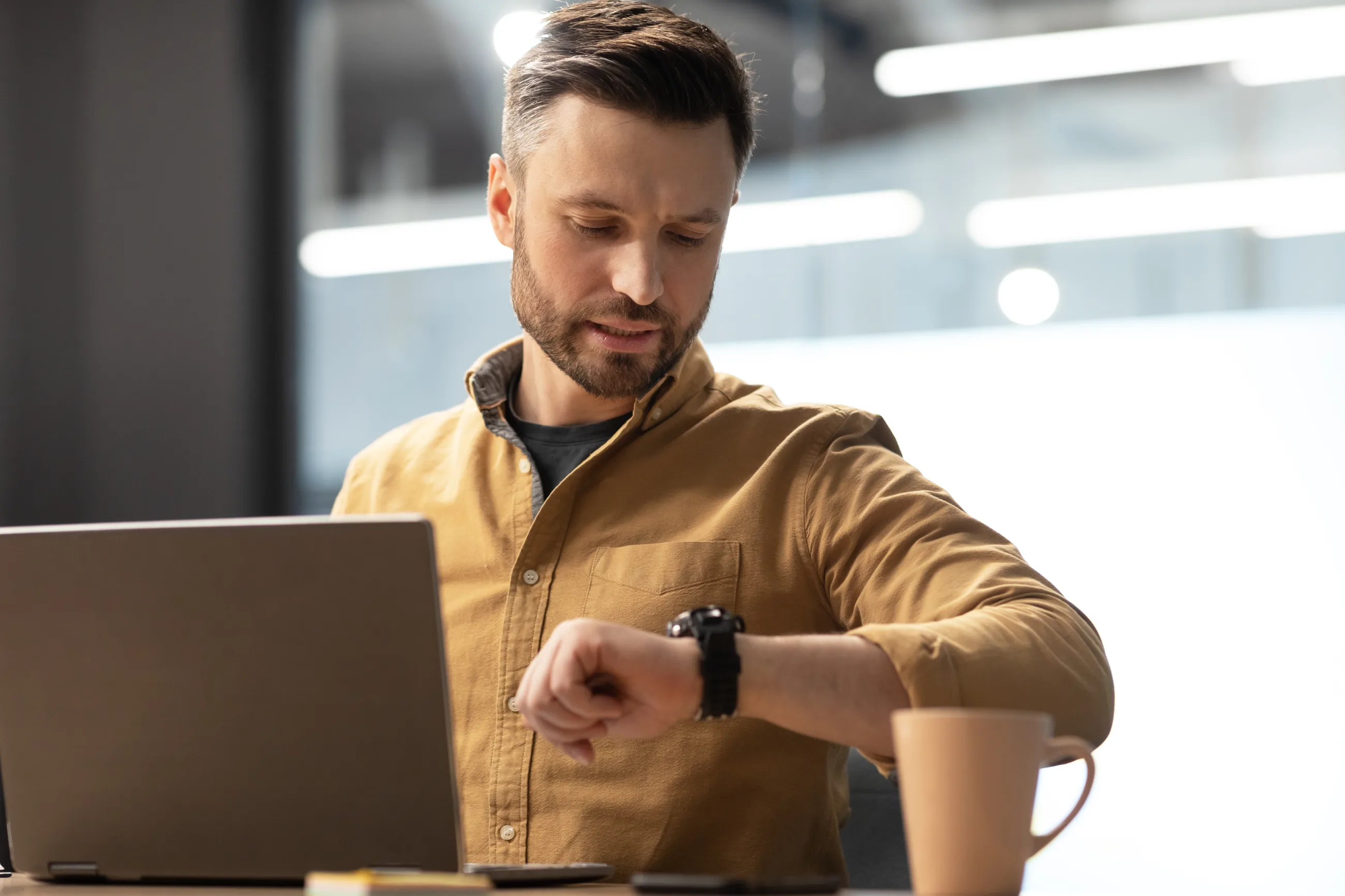 A man at his laptop checking his watch, a mug beside him